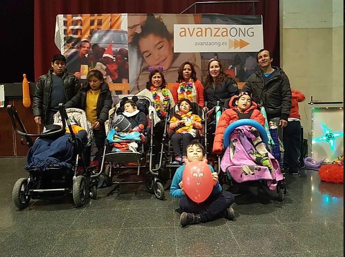 Familias y niños, algunos en sillas de ruedas o cochecitos, posan junto al cartel de avanzaONG y Un niño con globo rojo sentado en el suelo, todos abrigados