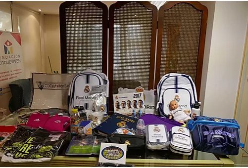 Sobre una mesa, productos del Real Madrid -mochilas, camisetas y más- apoyan la labor de cuidados pediátricos Fundación porqueViven. De fondo, un cartel de Fundación Enrique Yben muestra el compromiso con los niños y sus familias en Madrid