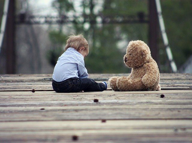 Un niño pequeño con camiseta azul y vaqueros, en un puente de madera junto a su osito, refleja el apoyo Fundación porqueViven