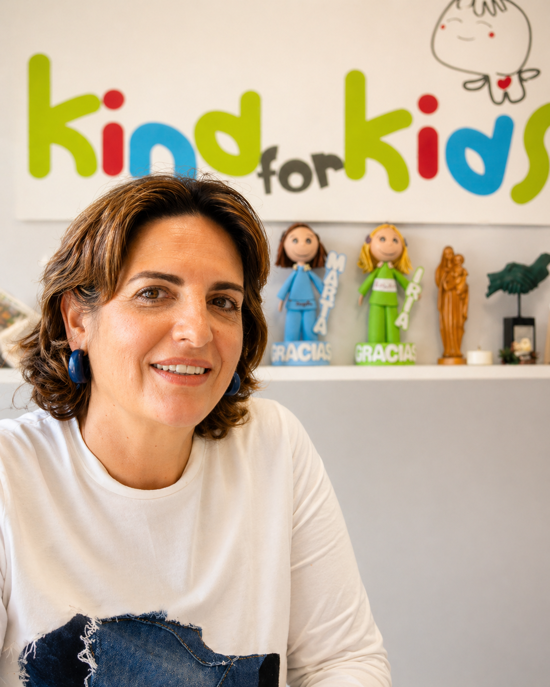 Una mujer con el pelo castaño hasta los hombros está sentada en un escritorio, sonriendo. Detrás de ella hay una estantería con figuritas y un cartel de "kind for kids" en la pared, que destaca el acompañamiento a niños con enfermedades graves. Lleva una camisa blanca con un parche azul.