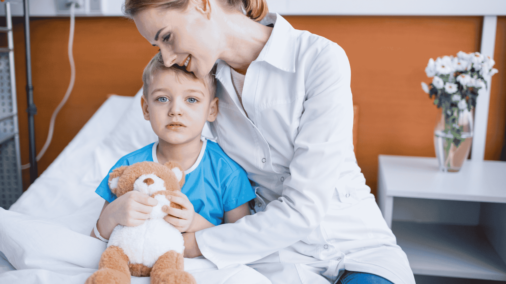 Un niño pequeño, en una cama de hospital, sostiene un osito de peluche, mientras una mujer con bata blanca —posiblemente una especialista de los Equipos de Atención Psicosocial (EAPS)— está sentada a su lado, inclinándose hacia él con un gesto reconfortante. Se ven flores sobre una mesa al fondo.